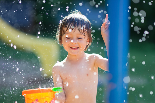 Cute little toddler boy, having fun with splashed water shot during summer time, holding bucket, outdoors