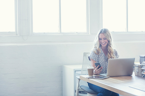 Attractive woman working on a laptop computer. She is casually dressed  with long blonde hair. She looks relaxed with a cup of coffee and she is probably surfing the internet. She could be a business woman working at home or in an office. Shot is back lit with copy space. There is a digital tablet and she is texting on a smart phone.