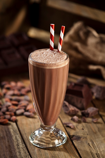 Classic glass of chocolate milkshake standing on rustic wood table. Chocolate chunks and cocoa beans are out of focus on the background. There are two old-fashioned red-and-white straws standing in the glass. Predominant color: brown.   DSRL studio photo taken with Canon EOS 5D Mk II and Canon EF 70-200mm f/2.8L IS II USM Telephoto Zoom Lens