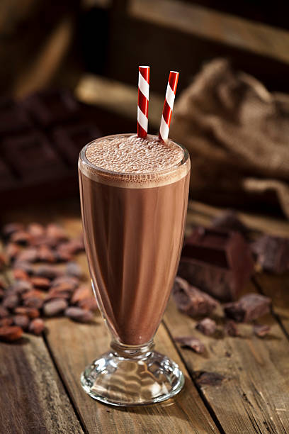 Chocolate milkshake on rustic wood table Classic glass of chocolate milkshake standing on rustic wood table. Chocolate chunks and cocoa beans are out of focus on the background. There are two old-fashioned red-and-white straws standing in the glass. Predominant color: brown.   DSRL studio photo taken with Canon EOS 5D Mk II and Canon EF 70-200mm f/2.8L IS II USM Telephoto Zoom Lens chocolate shake stock pictures, royalty-free photos & images