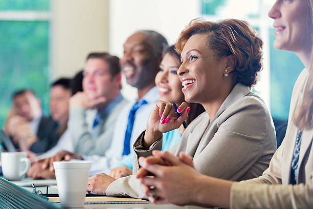 African American businesswoman attending seminar or job training business conference Mid adult African American businesswoman is smiling while attending a job seminar or job training business conference. She s taking notes while listening to a speaker. She is dressed in smart business casual clothing and is sitting next to diverse group of professional coworkers. corporate boardroom stock pictures, royalty-free photos & images