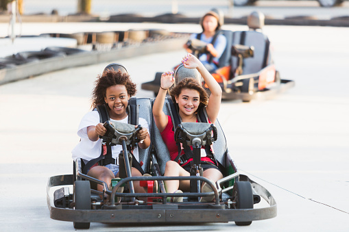 Multi-ethnic teenagers driving go carts at an amusement park.  Focus on teens in front, an African American girl and her Hispanic friend.