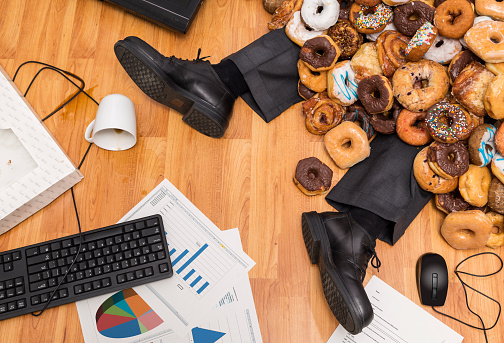 Overeating at work concept - A business person is buried under a large pile of donuts at their desk.  They have spilled coffee, reports on the floor, and the computer on the floor.  Please see my portfolio for other business concept images. 
