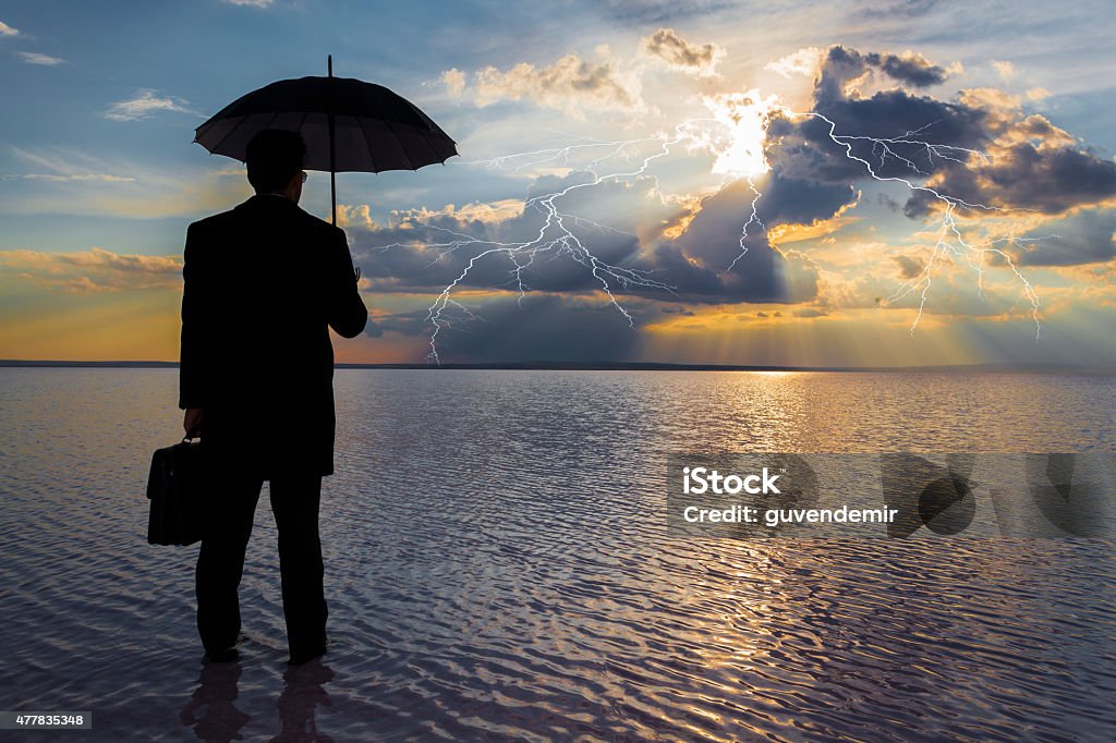 Businessman in trouble concept A rear view of a businessman in black suit with a briefcase and a black umbrella standing at water edge and looking rainstorm clouds at sunset. Lightning is seen descending from a gray and cloudy sky. Briefcase Stock Photo