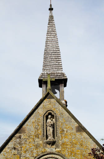 This is the chapel of St. Mary in Godstone, Surrey, southern England. The photograph shows a sculpture of Mary holding the baby Jesus, and features the chapel's crowning fleche and bell tower. (In English architecture, a fleche is a lead-covered timber spire, usually small in size.) The chapel was built in 1872-73 by Sir George Gilbert Scott as part of a scheme that included building Tudor-style almshouses for the poor. The building is Grade II listed, with the following description: (Chapel to left end: two storeys to front, one storey to rear with coved eaves to roof and shingled lead fleche over east end. Gabled end to street jettied on first floor with end brackets. Central angle bay to ground floor with leaded casement windows. Planked door to centre of south side (right) in gabled porch with date 1872 inscribed in gable. Interior:Two bay timber framed nave with stone fireplace to west end. Neo-Norman chancel to east with round chancel arch, mosiac inlay and wrought iron screen.).