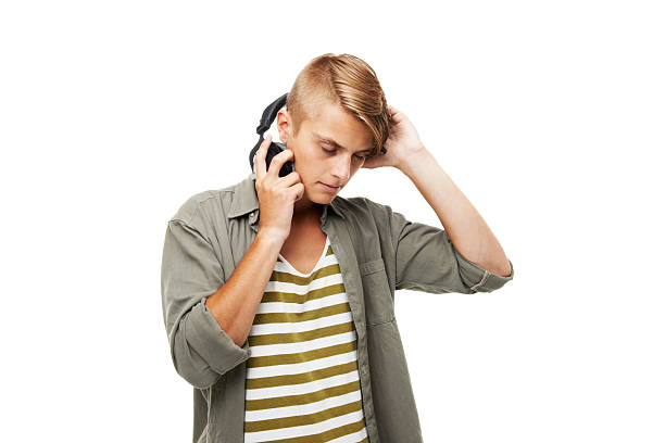 The smiling teenage boy or high schooler boy with headphones is holding mobile phone in hand outdoors.