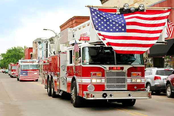 A group of fire trucks with American flags on them drive down the road in a small town American Parade during a festival event.