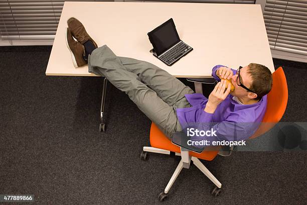 Man Sitting At The Desk Eating Hamburger In Office Stock Photo - Download Image Now