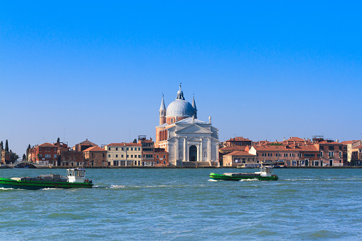 The imposing Chiesa del Santissimo Redentore (a Catholic church designed by Andrea Palladio and known as Il Redentore) in Venice, Italy is the focal point. The wide-angle shot also shows the waterfront of La Giudecca Island with colorful houses and palazzi.  A large swath of the Giudecca Canal runs along the bottom of the frame (with 2 green barges) and a brilliant blue sky at the top. Plenty of copy space available.