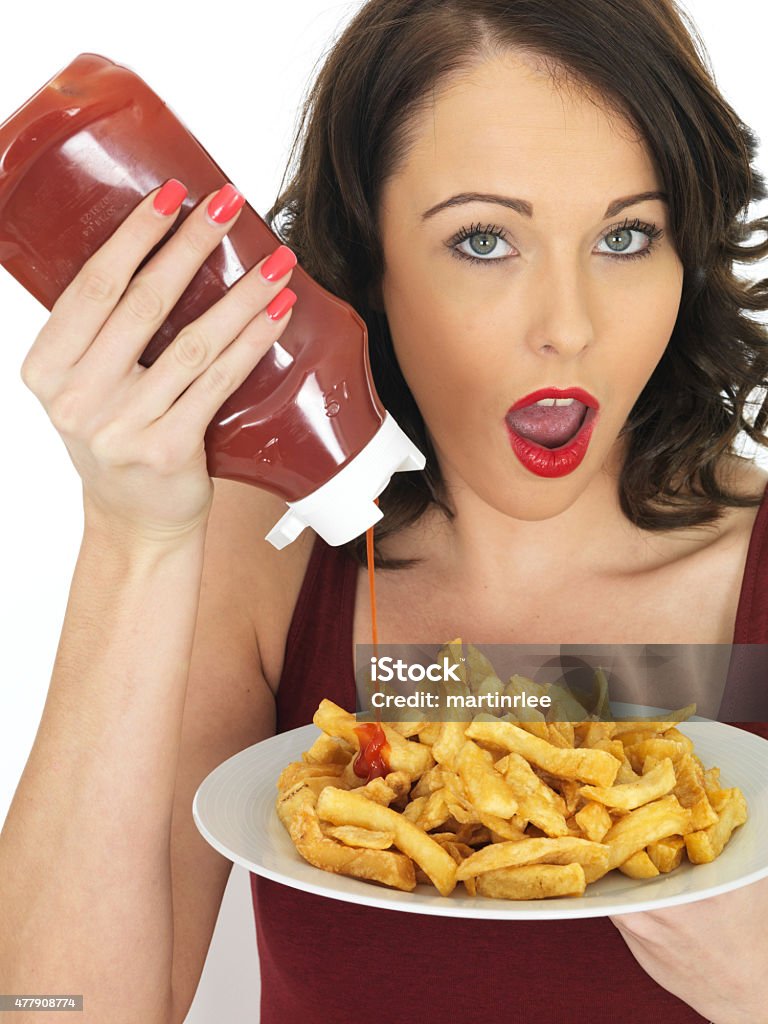 Young Unhealthy Woman Eating a Large Plate of Fried Chips Attractive unhealthy young woman, with dark mid length wavy hair,  holding a white large plate of fried takeaway chips holding a bottle of tomato ketchup squeezing the ketchup on top of the chips with shocked expression, wearing a casual dark red top, shot against a white background, looking into the camera 2015 Stock Photo