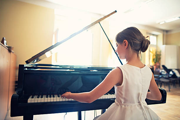 Little girl playing on grand piano Little girl playing grand piano in a sunny class room. She is preparing for a piano performance in music school. The girl aged 9 is sitting at the grand piano, back to the camera. The girl is smiling and wearing a white dress. girl playing piano stock pictures, royalty-free photos & images