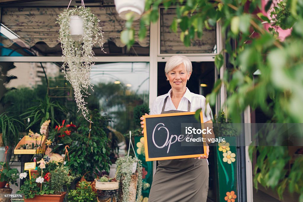 Smiling mature woman holding open sign at flower shop. Smiling mature woman holding open sign, written on blackboard with chalk, outside of flower shop. Looking at camera and smiling. Open Sign Stock Photo