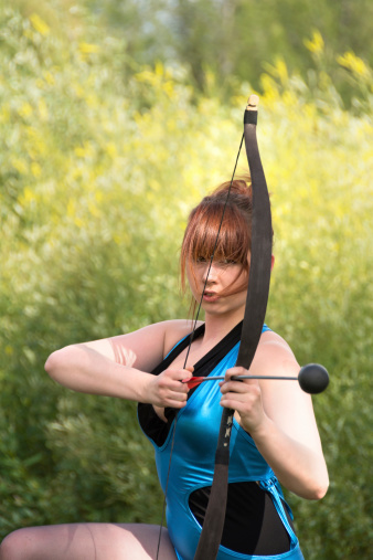 Vertical outdoor shot of young woman kneeling with bow and arrow considering next shot.
