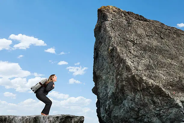 A businesswoman with a rocket strapped to her back preparing for liftoff in order to reach a higher level. She is standing on a cliff looking up towards a mountain top that she is trying to get to. The cliff and mountain top are rocky and barren.