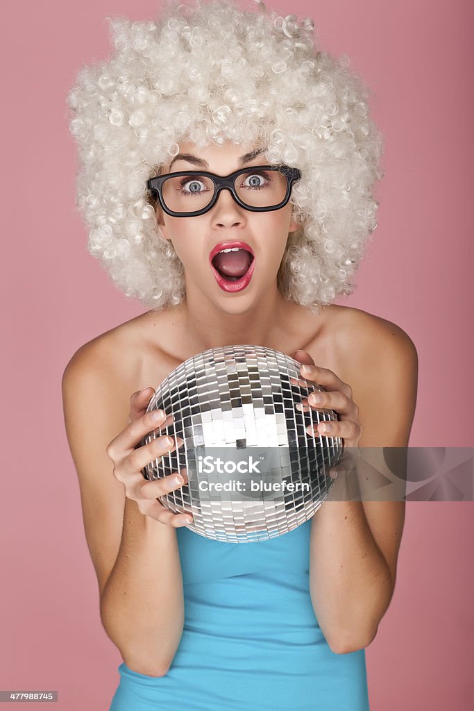 Having fun. Playful and funny woman wearing a curly wig on a pink background holding a disco ball. Novelty Glasses Stock Photo