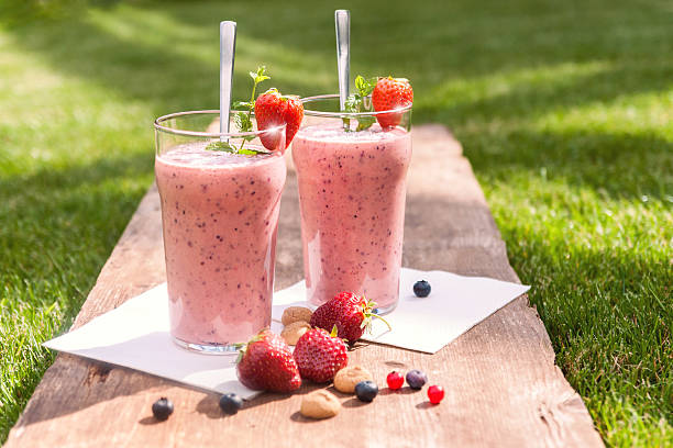 Fruity berry milkshake outdoors 2 glasses with a berry milkshake and decoration on an old wooden board lying on grass, landscape, direct sunlight yogurt fruit biscotti berry fruit stock pictures, royalty-free photos & images