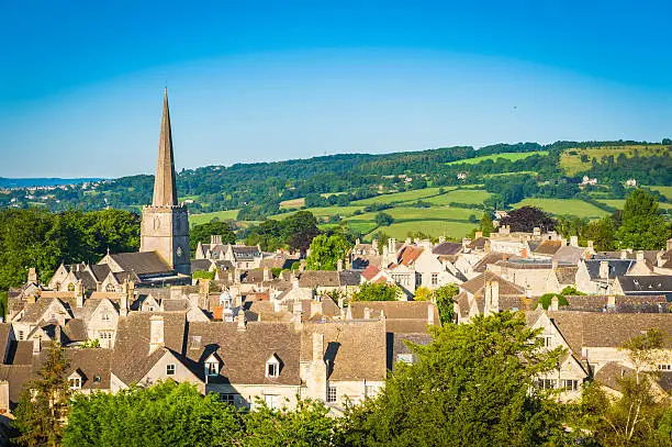 Aerial view over the iconic Cotswold village of Painswick, with its honey coloured limestone cottages and historic church spire, Gloucestershire, UK, framed by vibrant green patchwork fields and clear blue summer skies. ProPhoto RGB profile for maximum color fidelity and gamut.