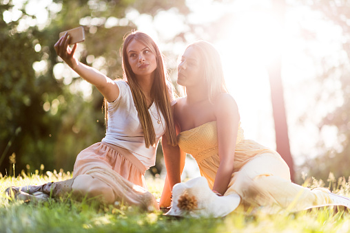 Young happy women relaxing in grass during spring day and taking a selfie with cell phone. They are puckering.
