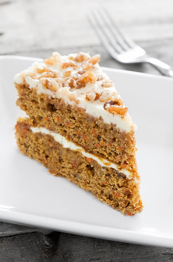 A slice of deliciously moist carrot cake is siting on a white ceramic plate. There is buttercream icing between the layers of orange sponge and on top of the cake which is decorated with chopped walnut pieces. The camera is selectively focused on the front of the cake.