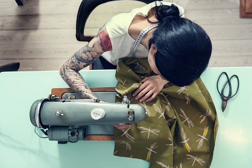 Overhead view of a female dressmaker working on a sewing machine in her design studio. Colour, horizontal format with a very shallow focus  point  on her hands as she works feeding the material through the machine. She is wearing a white sleeveless top that shows of her full sleeve tattoo she is half Japanese half european with dark hair.