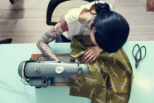 Professional Dressmaker At Work Overhead view of a female dressmaker working on a sewing machine in her design studio. Colour, horizontal format with a very shallow focus  point  on her hands as she works feeding the material through the machine. She is wearing a white sleeveless top that shows of her full sleeve tattoo she is half Japanese half european with dark hair. sewing item photos stock pictures, royalty-free photos & images