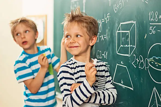 Cute little boys in math class, smiling back to the teacher. There are mathematic symbols written at the brackboard.