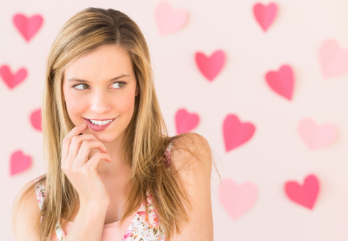 Shy young woman biting lip while looking away with heart shaped papers stuck against colored background