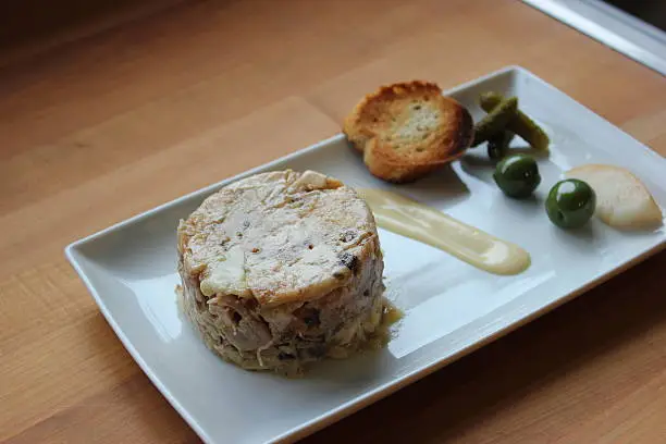 This image shows a cylinder-shaped chicken terrine served with pickles, crostini and an aoli over a white plate, on a butcher block and against a stainless steel backdrop. This is part 2 of a 3-part series, The other images show different angles (up top, from the side, etc.)