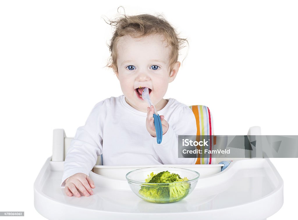 Cute funny baby with big beautiful blue eyes eating broccoli Cute funny baby with big beautiful blue eyes eating broccoli in a white high chair, isolated on white Baby - Human Age Stock Photo