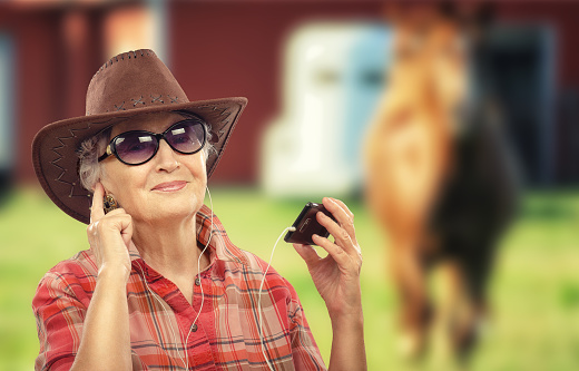 Aged cowgirl is listening country music with smart phone. She is wearing brown cowboy hat and red plaid shirt. Old cowgirl in earphones is posing with sunglasses. There is a horse on the background out of focus