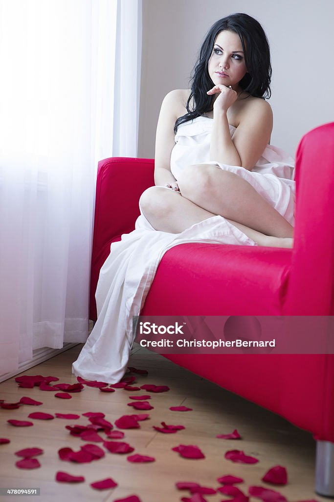 Bella donna seduta sul divano rosso con petali di rosa - Foto stock royalty-free di 20-24 anni