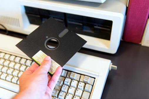 Old way of working with computers: man is holding a floppy disk which is being inserted into a on old pc.