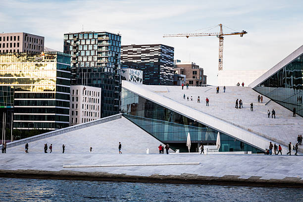 Oslo, Norway - June 12, 2015: the modern architecture of the Oslo Opera House in Oslo, Norway. Tourists walk on the marbled paving that surrounds the opera house. In the background is Oslo's modern business district, made up of multiple modern skyscrapers.