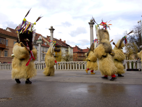 Ljubljana, Slovenia -March 1, 2014: Blurred Kurents, with traditional fur masks, dancing on Ljubljana bridge during carnival. In background is  bridge fence and surrounding building with cloudy sky above them.