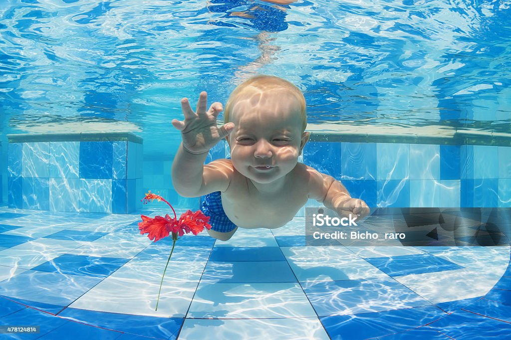 Child swimming underwater for a red flower in the pool Smiling baby boy diving underwater with fun for red flower in blue pool Active lifestyle, child swimming lesson with parents, and water sports activity during family summer vacation in tropical resort Baby - Human Age Stock Photo