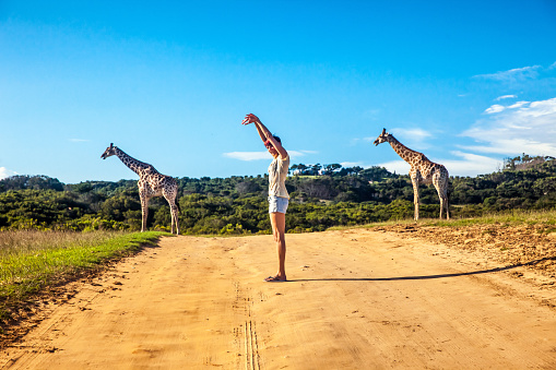 Woman standing in the  middle of a dirt road in Africa making a fun posture to resamble the two giraffe in the background.