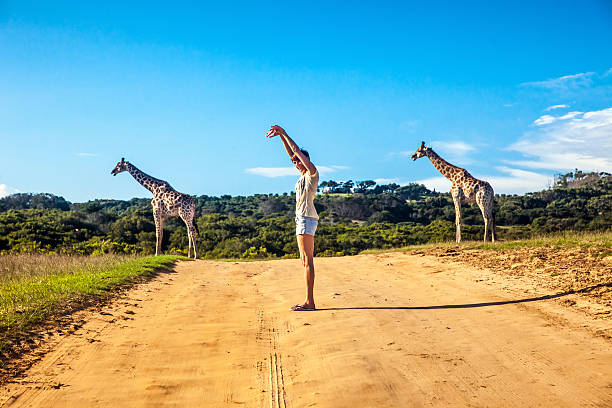 Giraffe Woman standing in the  middle of a dirt road in Africa making a fun posture to resamble the two giraffe in the background. giraffe photos stock pictures, royalty-free photos & images