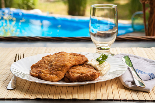 Breaded Poultry fillet served outdoor by a swimming pool on white plate with Mayonnaise decorated with few leaves of parsley. Close up with visible cross section of white meat. Empty glass in blurred background.