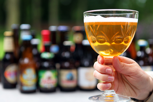 A person is holding a full glass of Belgian abbey beer in her hands. Variety of Belgian beer bottles are seen blur in the background, providing space for your text.