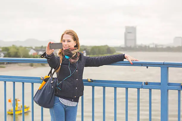 German girl taking selfie in Bonn with Rhein on background. She is standing next to the railing, holding the smart phone with one hand and looking at it.