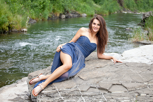 Young attractive brunette in long blue dress is lying on a rock by the river. Blurred background of the river and green nature, she is smiling and looking at the camera. Summer atmosphere. Shooting with Canon EOS 5D Mark II.