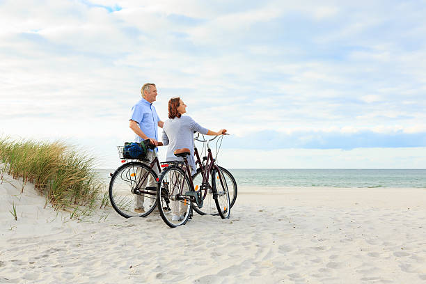 matura coppia all'aperto sulla spiaggia - mature adult bicycle senior adult heterosexual couple foto e immagini stock