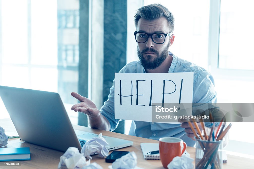 Asking for help. Frustrated young beard man holding piece of paper and asking for help while sitting at his working place in office 2015 Stock Photo