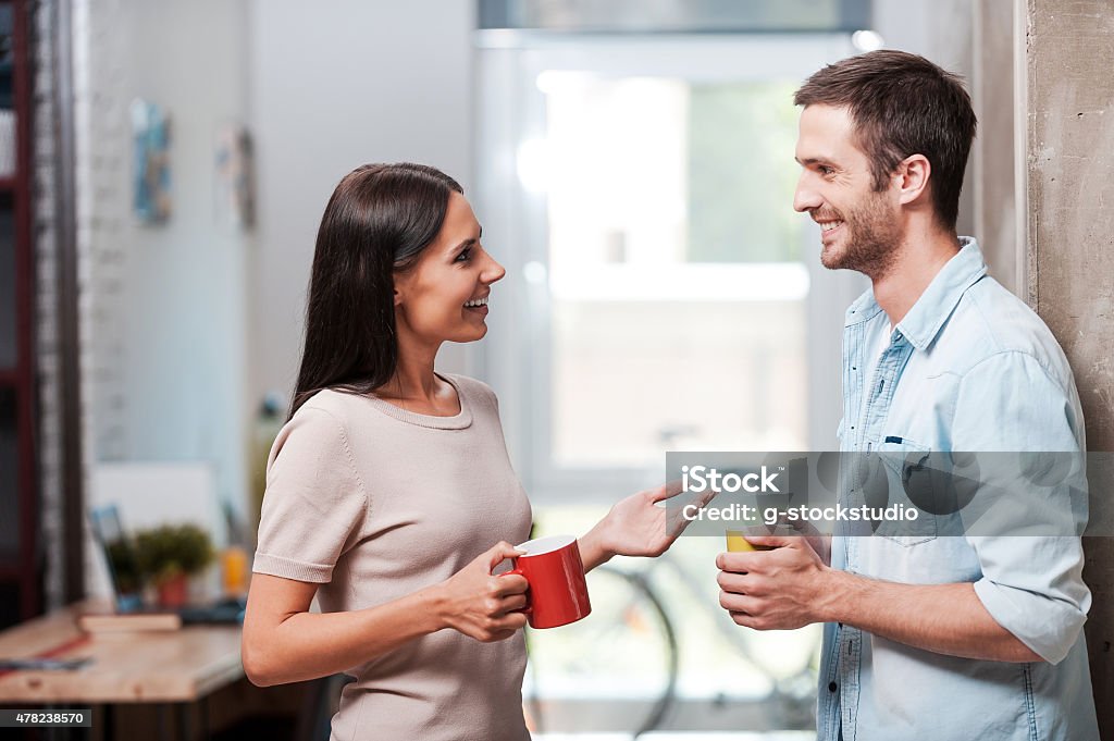 Spending a nice coffee break. Two cheerful young people holding coffee cups and talking while standing in office Discussion Stock Photo
