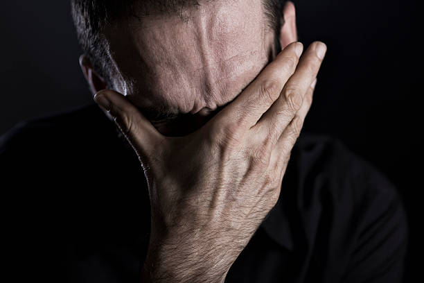 Close up of depressed and despaired man. Close up of miserable man burying face in hans, looking desperate, isolated on black background. head in hands stock pictures, royalty-free photos & images