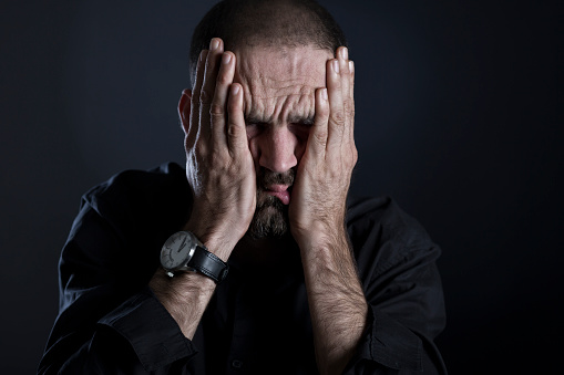 Stressed man looking exhausted, covering face with hands, isolated on black background.