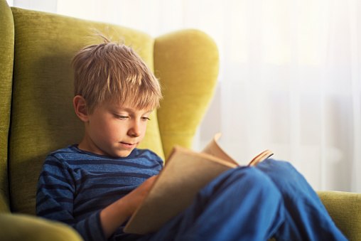 Little boy sitting on a gree armchair near the window, reading a book. The boy is aged 5 and is wearing blue clothes.