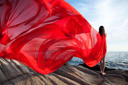 Beautiful young woman wearing red dress and standing on the rocks on coast by the sea while wind blowing in the dress. Her dress swinging in foreground and makes wonderful pattern with some perts blurred in motion. Girl is looking at horizon and looks lonely