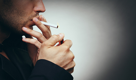 Closeup of unrecognizable caucasian man lighting a cigarette. he's wearing black coat and has short beard. Shot over vignetted gray background.