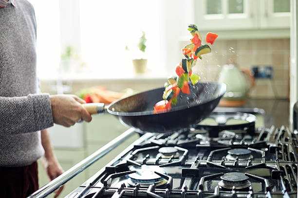 Man tossing fresh vegetables in saucepan at kitchen Midsection image of man tossing fresh vegetables in frying pan. Various vegetable slices are in mid-air. Male is preparing food in domestic kitchen. camping stove photos stock pictures, royalty-free photos & images